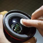 Proper lens cleaning method showing microfiber cloth or cleaning tool being used on camera lens glass to remove dust and smudges safely