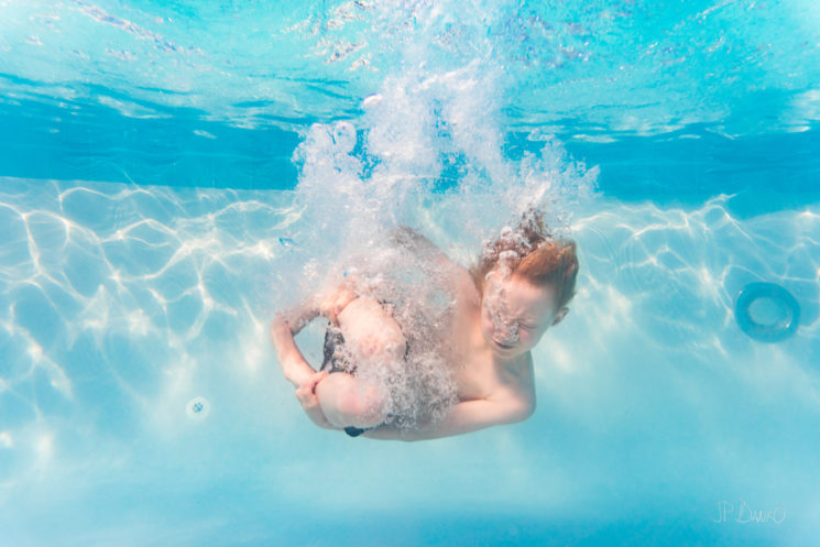 Underwater view of redhaired boy diving cannonball in cool blue holiday swimming pool