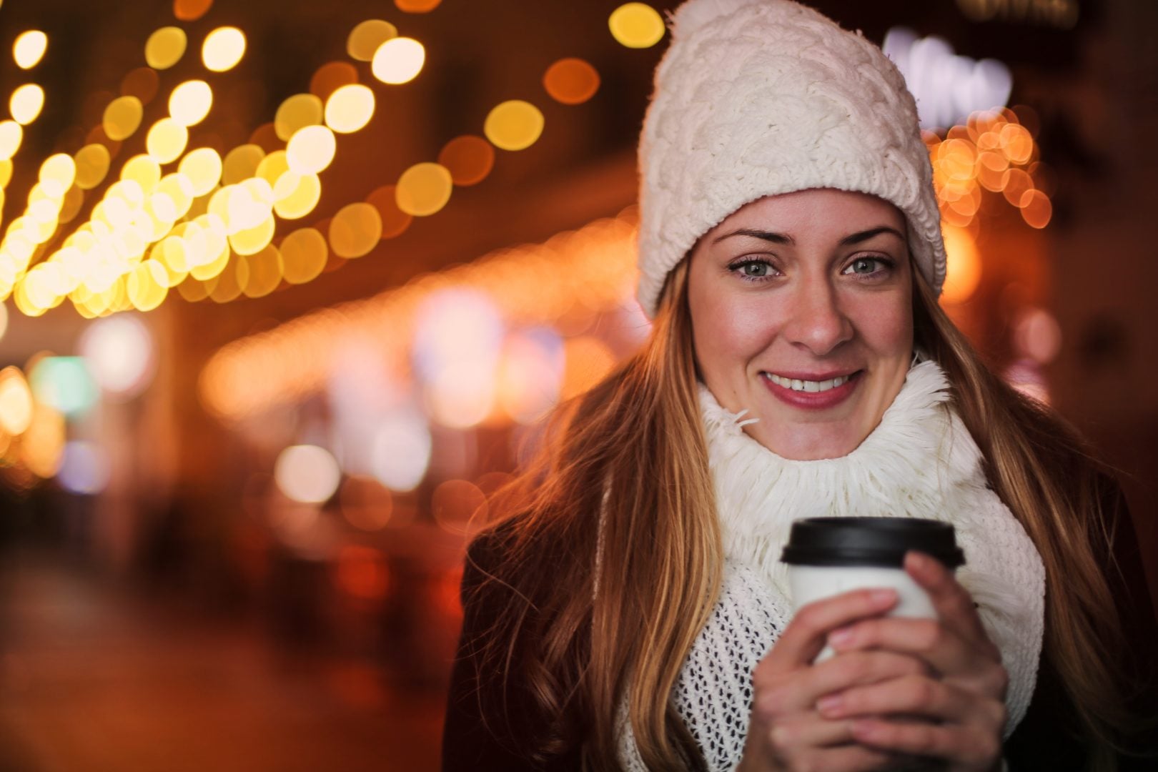 Woman in a white knitted hat and scarf, holding a takeaway hot drink with bright lights behind her.