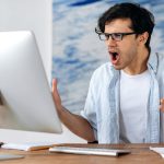 Outraged irritated young hispanic man with glasses, broker, businessman or manager, got financial loss, breakdown of the deal, displeased looks at the computer screen, gesturing with his hands