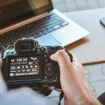 Close-up of hands holding DSLR camera at a workspace with laptop and smartphone.