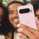 A person holding a pink Google Pixel smartphone while taking a selfie with two smiling friends in the background.