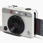 The Leica x Moncler Sofort 2 instant camera in white, featuring a sleek design with Moncler's logo and a black lens, printing an instant photo.