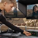A filmmaker adjusts a Blackmagic setup on a mobile workstation, with dual monitors displaying a model in a desert scene