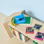 A bright blue Polaroid Gen 3 instant camera with "MoMA" branding sits on a wooden shelf beside colorful photo prints and books, showcasing a collaboration between Polaroid and the Museum of Modern Art.