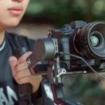A young man holding a camera mounted on a gimbal stabilizer, filming with a TTArtisan 35mm T2.1 cine lens in an outdoor setting.