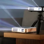 Two compact Kodak Luma 500 Google TV projectors on a table, one mounted on a small tripod, both projecting bright beams in a dimly lit room.