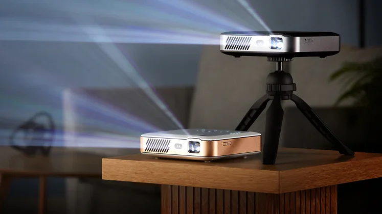 Two compact Kodak Luma 500 Google TV projectors on a table, one mounted on a small tripod, both projecting bright beams in a dimly lit room.