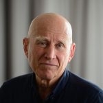 Portrait of Sebastião Salgado with a slight smile, wearing a dark shirt, standing in front of a softly blurred background with vertical lines.