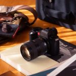 Leica SL mirrorless camera with a Vario-Elmarit-SL 28–70mm f/2.8 lens placed on a book, surrounded by travel accessories like a bag, sunglasses, and LFI magazines on a wooden table.
