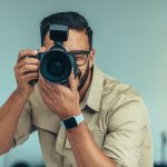 A portrait photographer in a beige shirt capturing a subject with a DSLR camera equipped with a flash, perfectly illustrating a professional studio setup ideal for sharp, well-lit portrait photography in 2025.