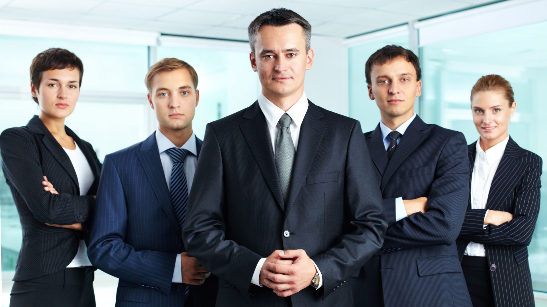 A professional corporate team of five individuals dressed in formal business attire, posing confidently in a modern office environment.