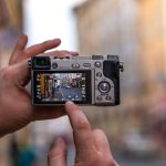 A close-up shot of a person holding a compact mirrorless camera, adjusting settings on the LCD screen while photographing a cobbled street.