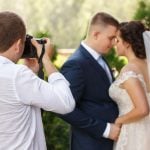 A photographer taking a shot of a couple on their wedding day, with a blurred background of greenery, capturing an intimate moment between the bride and groom.