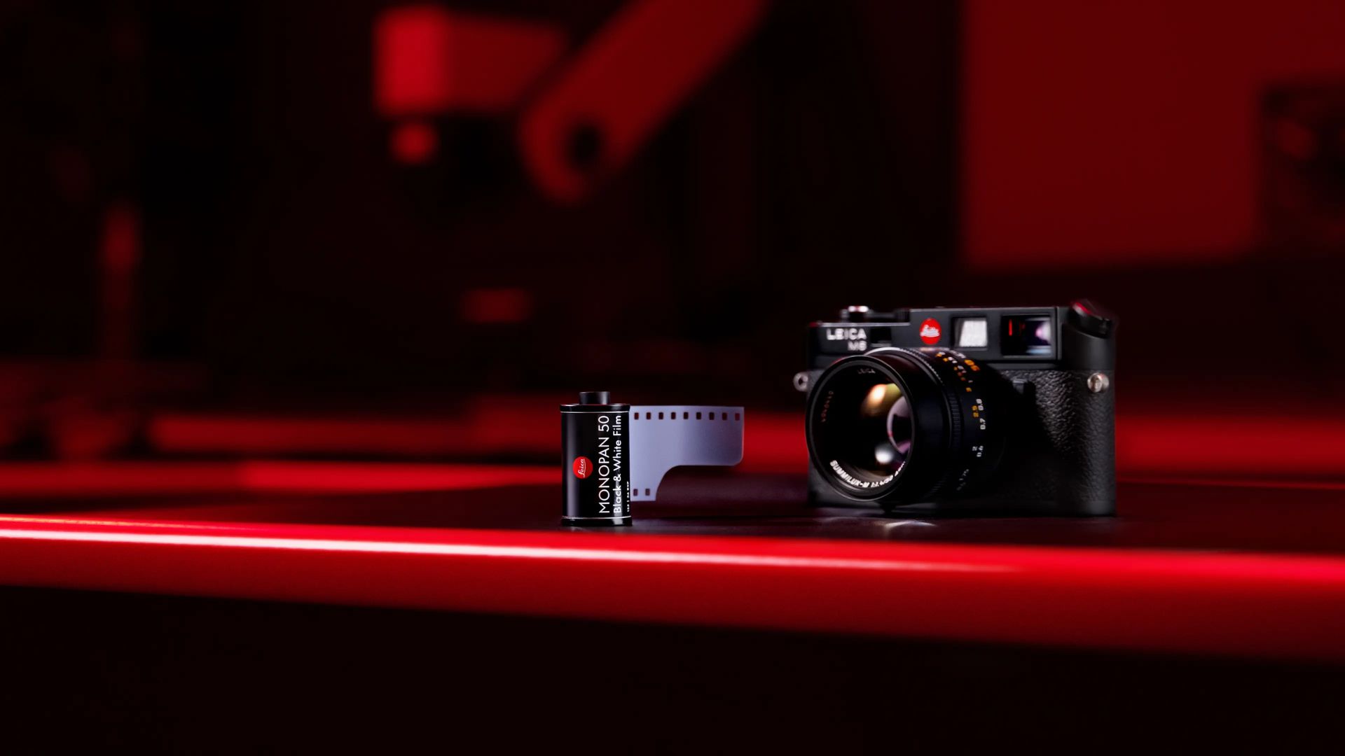 A Leica M-series camera with a black and white Leica Monopan 50 film roll placed beside it, all bathed in soft red lighting, showcasing the classic and modern fusion in photography gear.