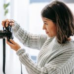 A woman in a cozy sweater holding a camera and adjusting its settings, seemingly in a well-lit indoor environment, focusing intently on the camera's display screen.