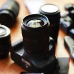 Black DSLR camera with attached lens surrounded by multiple camera lenses and photography equipment arranged on wooden surface in warm lighting.