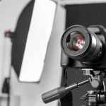 Black DSLR camera with telephoto lens mounted on tripod in monochrome studio setting with softbox lighting equipment visible in background.