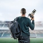 Professional photographer with camera backpack and telephoto lens standing on stadium field, facing empty grandstands under dramatic sky lighting.