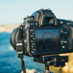 Close-up of a DSLR camera mounted on a tripod, capturing a scenic ocean view with rocky cliffs in the background — a timeless choice for photography enthusiasts.