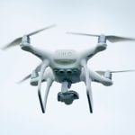 Modern white drone with four spinning rotors and mounted camera hovering in flight against bright overcast sky during aerial photography session.