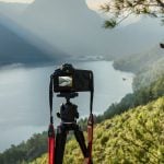 Professional DSLR camera mounted on tripod positioned to photograph scenic mountain lake view with forested hills and pine tree foreground.