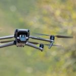 Gray DJI Mavic 3 quadcopter drone hovering mid-flight with motion-blurred propellers against soft-focused green foliage background during outdoor aerial photography.