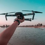 Person's hand holds compact DJI Mavic Pro drone above calm lake water during golden hour with pink sunset sky and desert landscape.