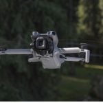 White DJI Mini 5 Pro quadcopter drone with large camera gimbal hovering in flight against blurred green forest background with spinning propellers.