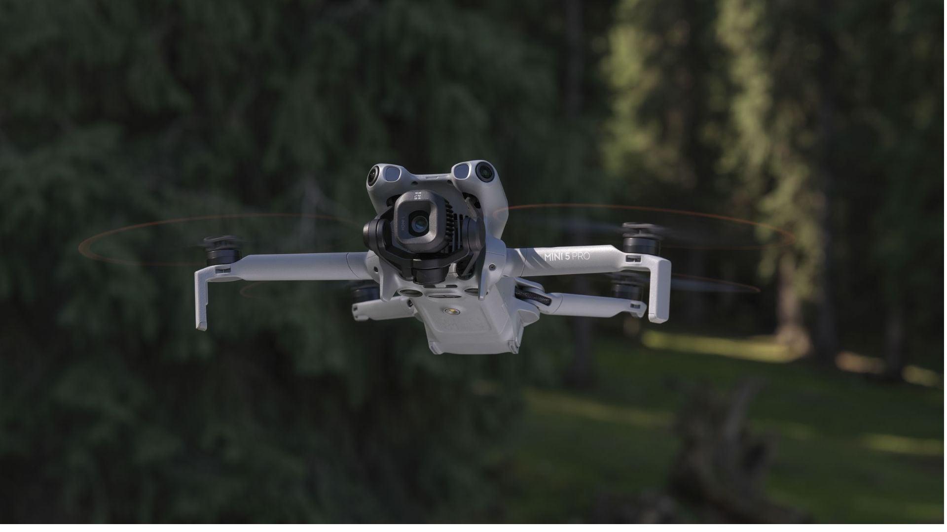 White DJI Mini 5 Pro quadcopter drone with large camera gimbal hovering in flight against blurred green forest background with spinning propellers.