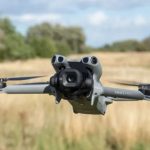 White DJI Mini 5 Pro quadcopter drone with large camera gimbal hovering above golden wheat field with trees and blue sky background.