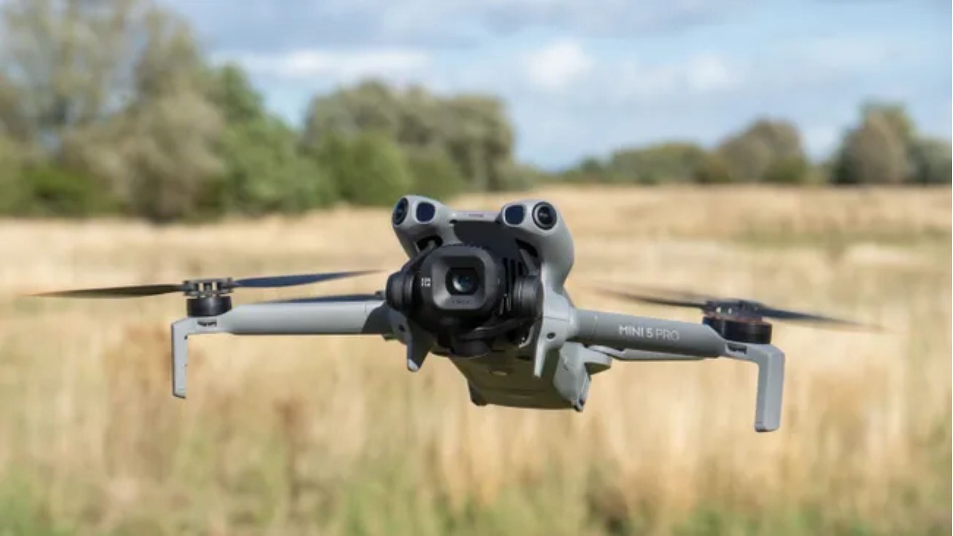 White DJI Mini 5 Pro quadcopter drone with large camera gimbal hovering above golden wheat field with trees and blue sky background.