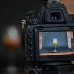 Black professional camera mounted on tripod showing colorful wine glass photograph on rear LCD display during studio product photography session.