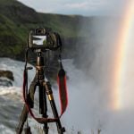 Professional camera mounted on tripod with LCD screen displaying waterfall photograph, positioned near actual waterfall with rainbow visible in misty background.