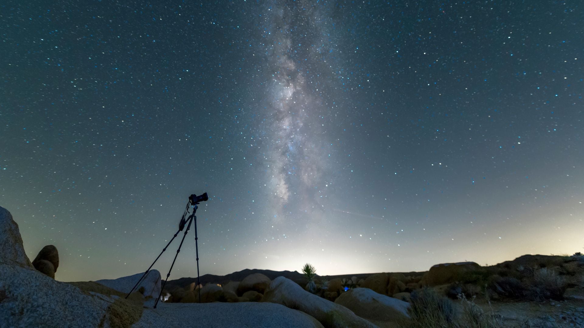Professional camera mounted on tripod positioned among desert rocks, capturing stunning Milky Way galaxy stretching across star-filled night sky.