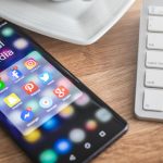 A close-up view of a modern Android smartphone displaying various social media applications such as WhatsApp, Facebook, Instagram, Pinterest, and Twitter, placed next to a white keyboard on a wooden desk — highlighting the phone’s everyday utility for sharing high-quality photos.