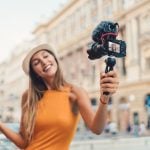 A woman holding a camera with a microphone attachment, smiling as she vlogs outdoors with a blurred city background.