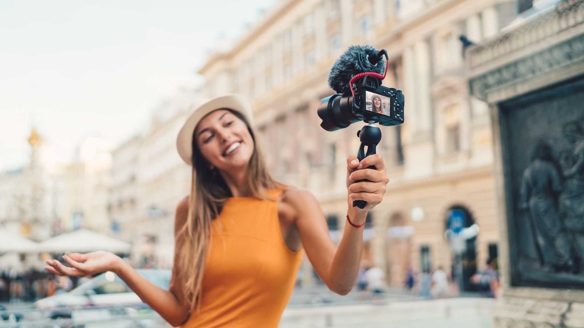 A woman holding a camera with a microphone attachment, smiling as she vlogs outdoors with a blurred city background.