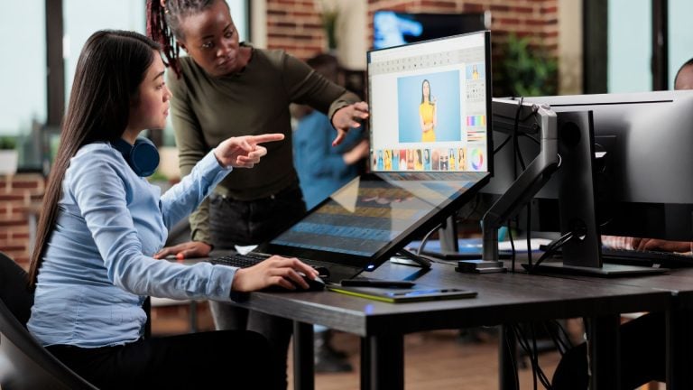 Two women collaborating on photo editing at a desk, working on a photo of a model on a large screen, demonstrating teamwork in the creative process with free photo editing software.