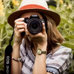 A woman with a straw hat takes a portrait photograph with a Canon DSLR camera in a sunflower field, emphasizing the shift from smartphones to professional cameras for budding photographers.