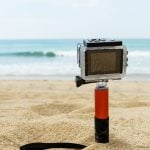 A GoPro action camera mounted on a stick is placed on the sand at the beach, with the ocean and sky in the background. The camera is ready to capture action-packed moments in an outdoor environment.