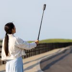 A woman is standing outdoors with a long selfie stick, holding a 360 camera to capture a wide scenic shot along a paved path surrounded by greenery.