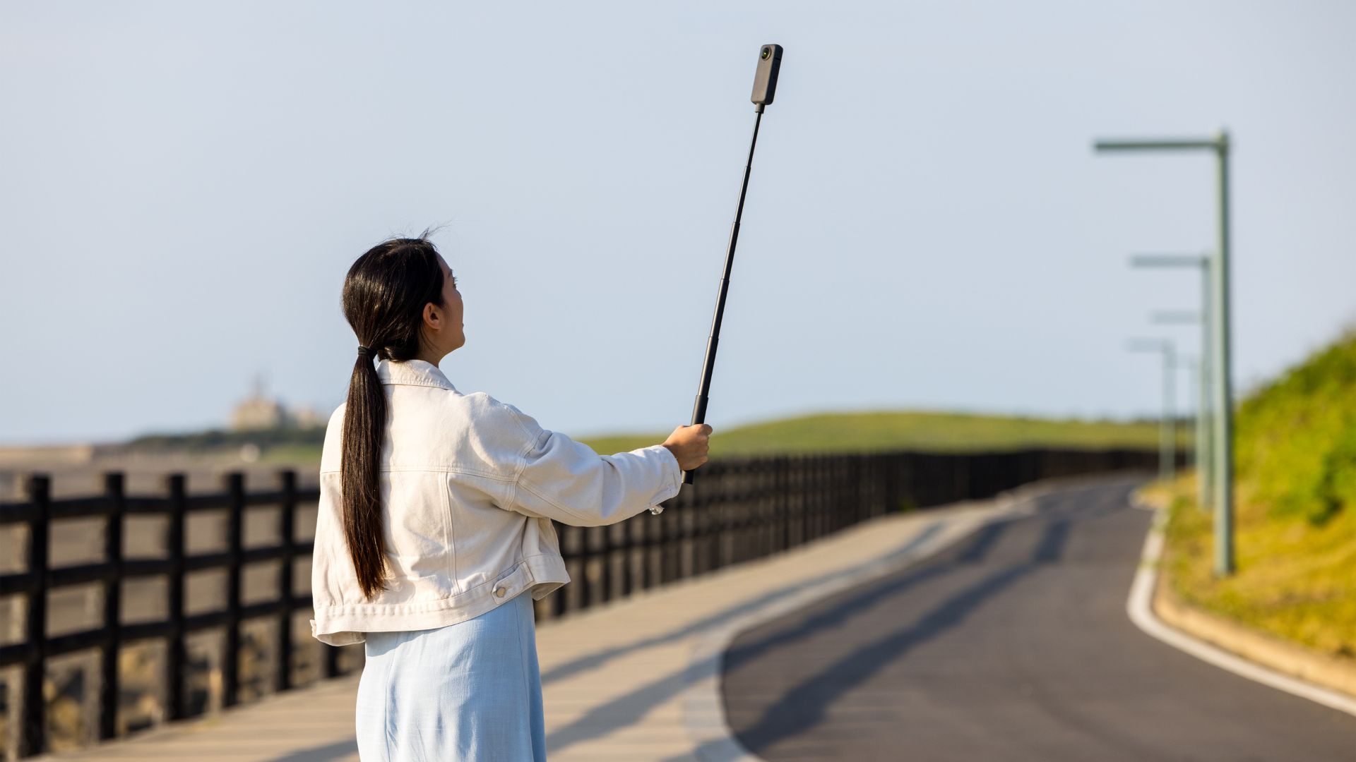A woman is standing outdoors with a long selfie stick, holding a 360 camera to capture a wide scenic shot along a paved path surrounded by greenery.