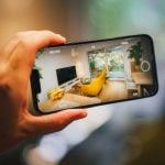 A person holding a smartphone, capturing a photo of a modern living room with visible manual camera settings like ISO and shutter speed displayed on the screen.