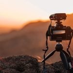 DSLR camera equipped with external microphone attachment on tripod, positioned on rocky outcrop overlooking layered mountain ranges during golden sunset.
