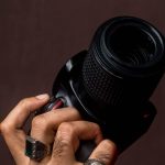 A close-up image of a person holding a professional DSLR camera by its lens. The camera’s lens is being carefully adjusted, with focus on the intricate design of the lens and the smooth handling of the gear.