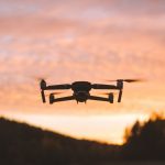 Black drone silhouette hovering against dramatic orange and pink sunset clouds with rolling hills and forest landscape below.