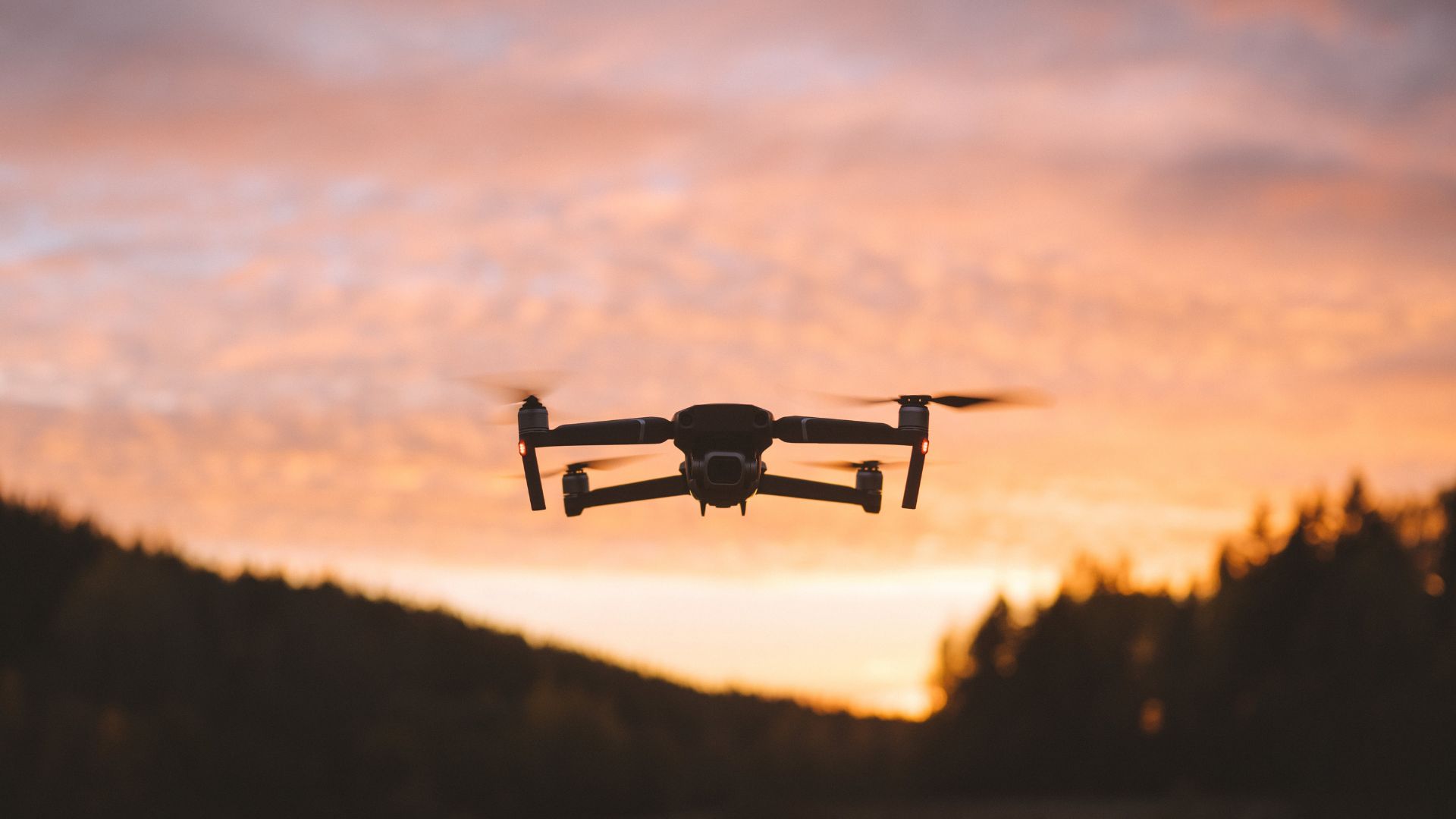 Black drone silhouette hovering against dramatic orange and pink sunset clouds with rolling hills and forest landscape below.