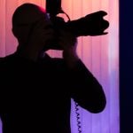 Dark silhouette of photographer holding camera to face against dramatic purple and pink colored studio backdrop with white curtain during photo session.