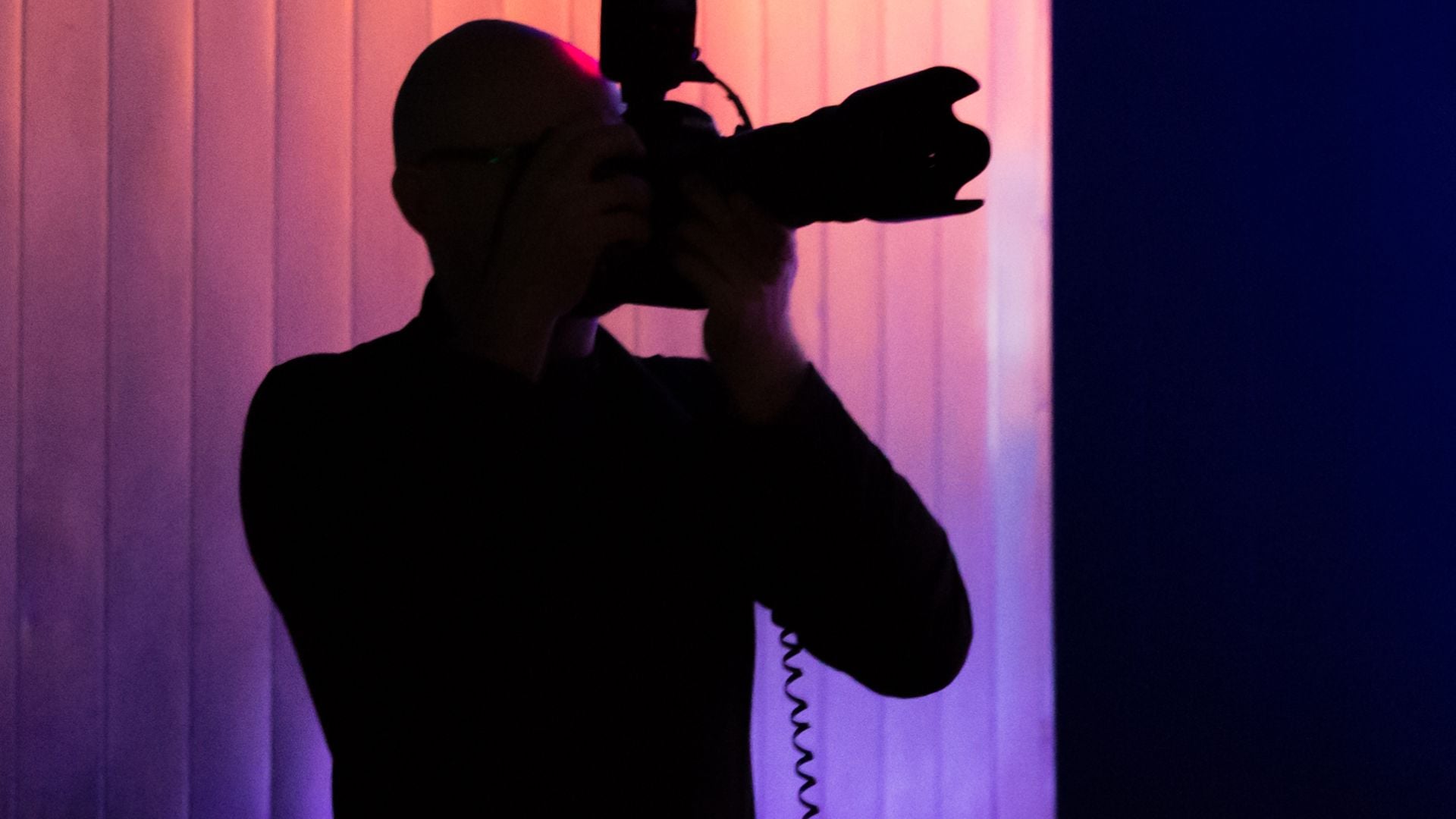 Dark silhouette of photographer holding camera to face against dramatic purple and pink colored studio backdrop with white curtain during photo session.